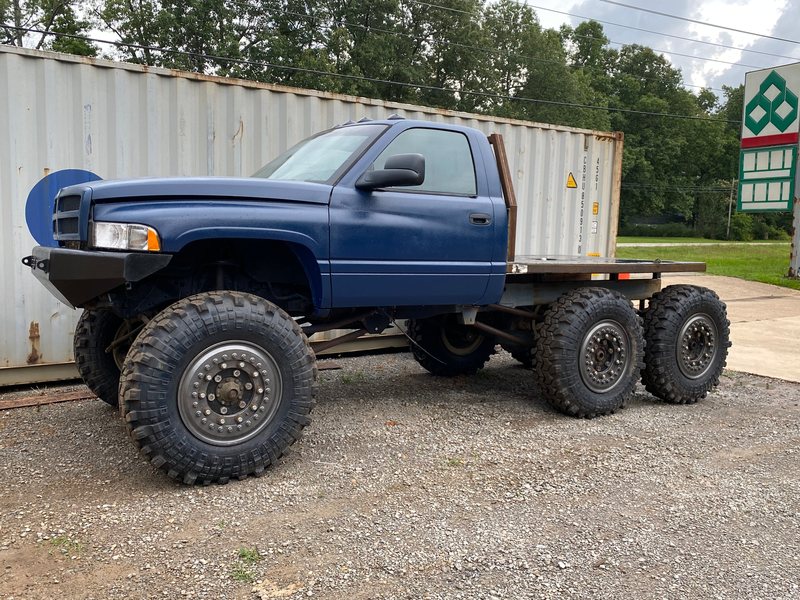 Side three-quarter view of the blue Ram 6x6 with its steel flatbed and headache rack, parked on gravel in front of a shipping container